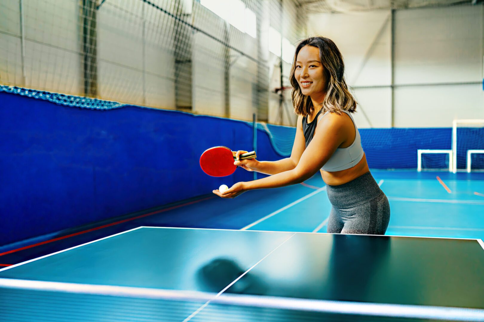 A smiling athletic woman is captured in an indoor sports setting, playing table tennis with a paddle in hand, ready to strike the ball