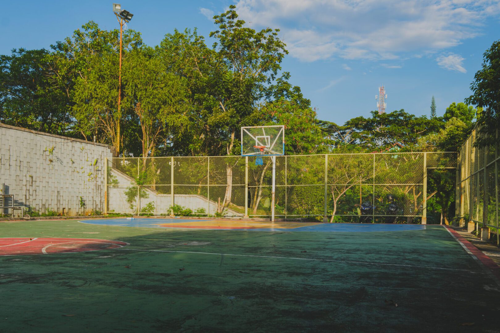 A basketball court bathed in warm sunlight, with a basketball hoop and a chain-link fence surrounding the court. The surrounding trees create a lush, natural setting.