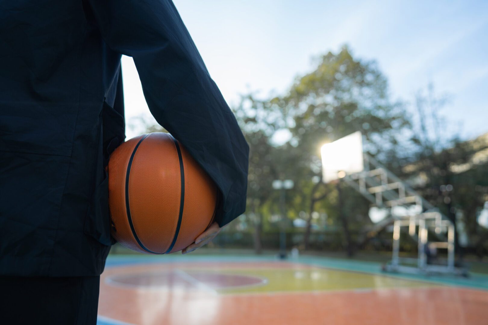 A young man strolling past the outdoor court, heading home after an enjoyable game, carrying a gym bag over his shoulder and holding a basketball in his other hand, showing positivity and a sense of calm relaxation