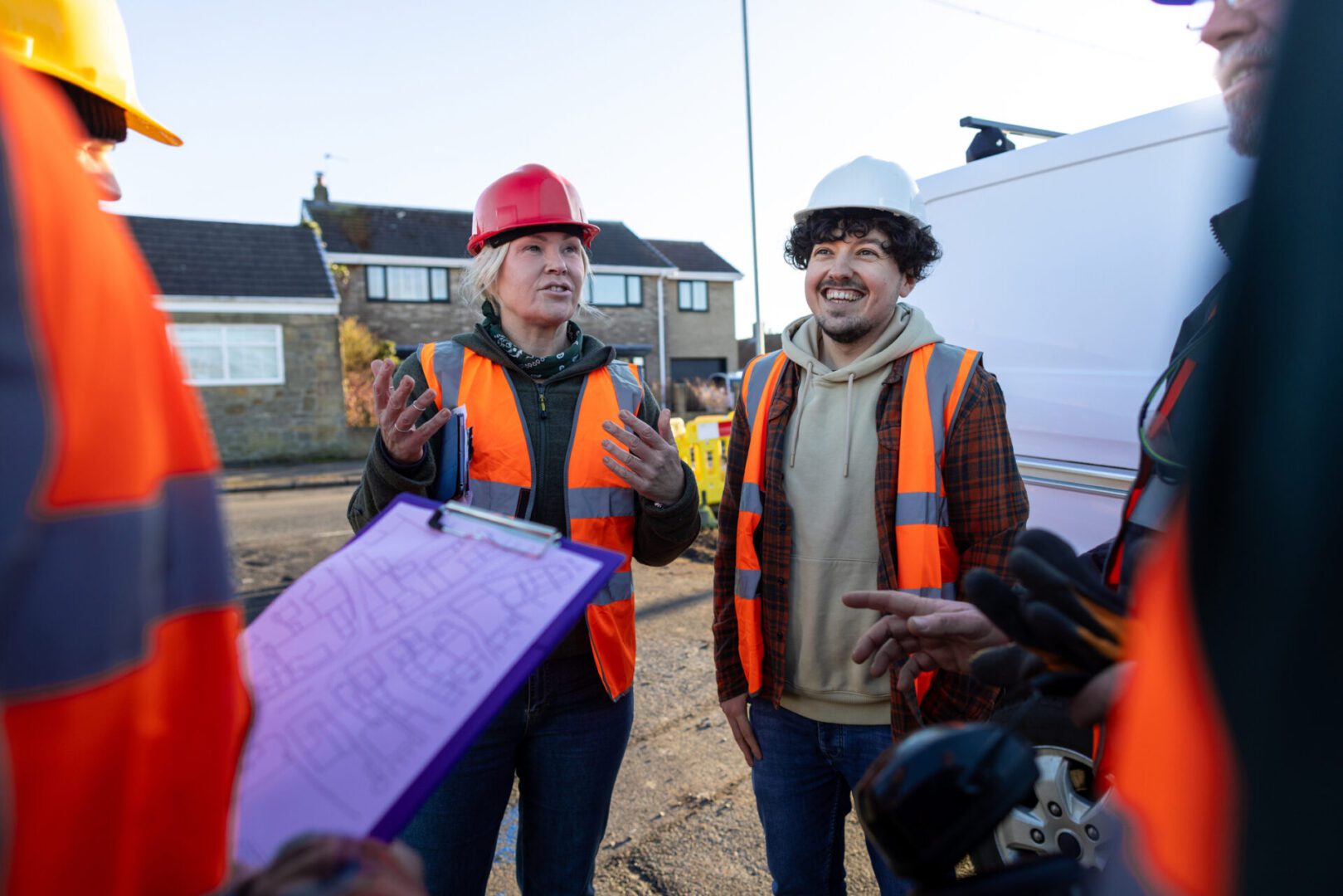 Front-view of a small group of construction workers engaged in a discussion at an outdoor worksite in Seghill, North East England. They're wearing safety gear, including hard hats and high-visibility vests, while standing together on site near construction barriers and their work van. One construction worker is holding blueprints of the street.

Videos similar to this scenario are available.