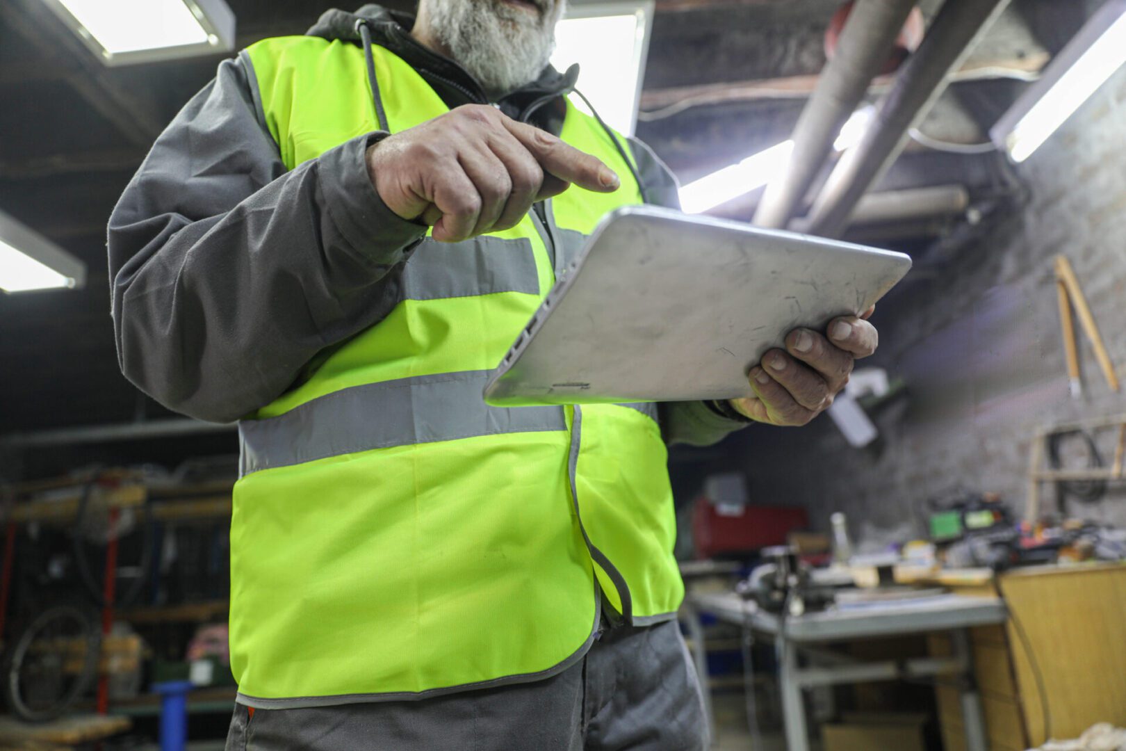 Older male in industrial attire using a tablet computer to manage maintenance or inspection work in a workshop. Concept of efficient construction management with technology.