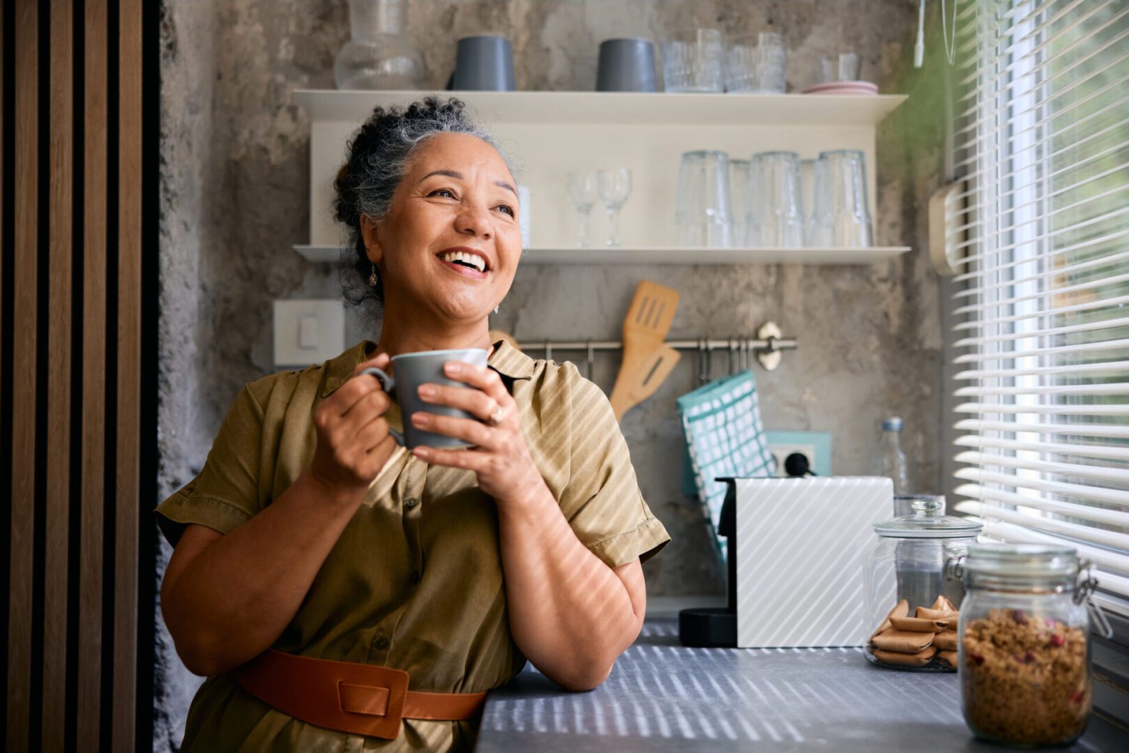 Cheerful mature woman sipping coffee in a modern kitchen, smiling while gazing out the window, embracing the peaceful morning and enjoying a moment of tranquility and relaxation