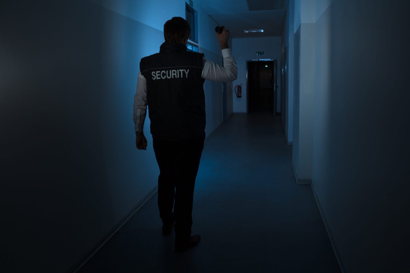 Rear View Of A Security Guard Standing In Corridor Of The Building Using Flashlight